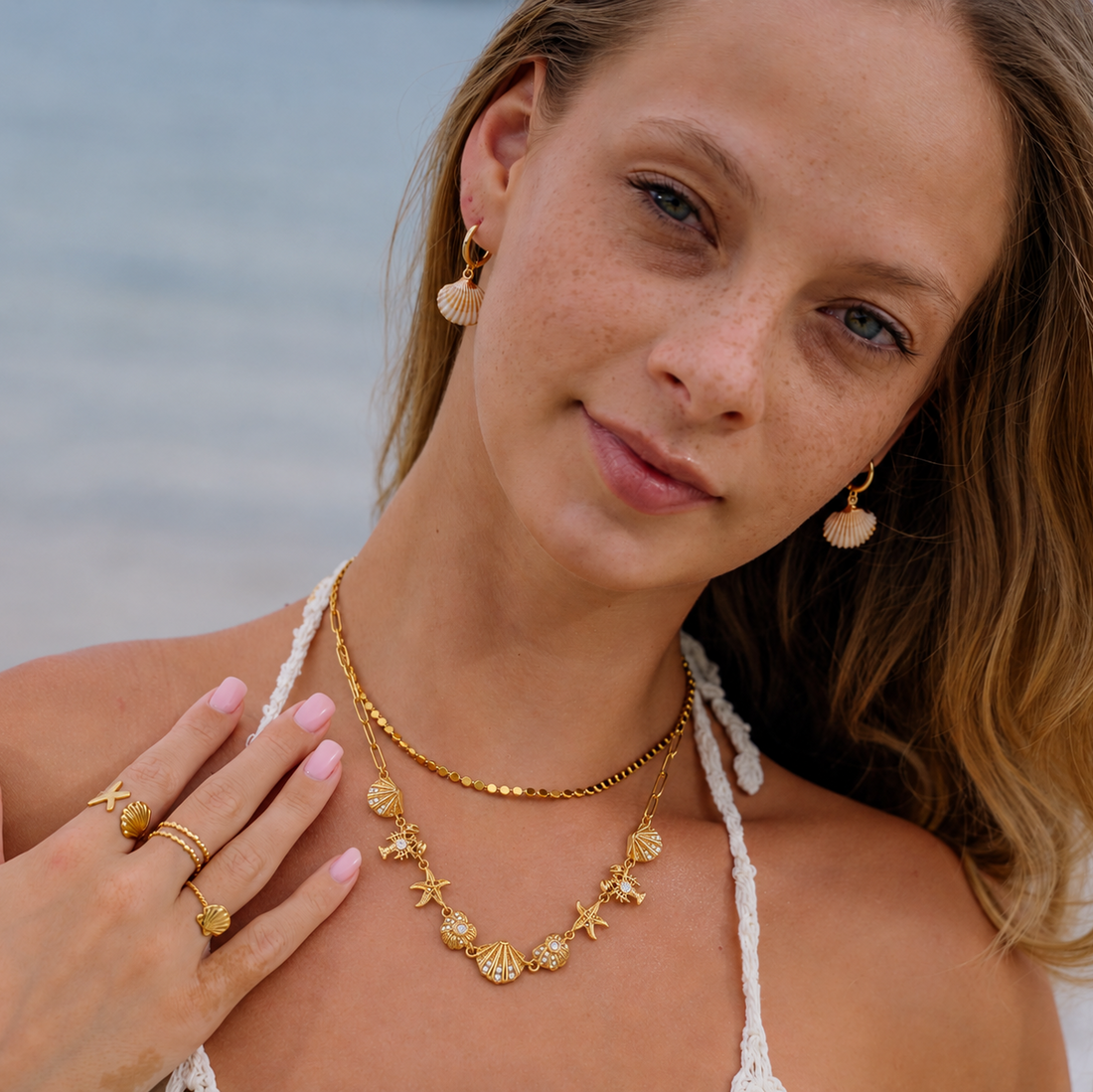 A close-up lifestyle shot of a woman at the beach wearing a gold necklace with starfish and seashell charms, matching starfish and seashell earrings, and gold rings on her fingers.