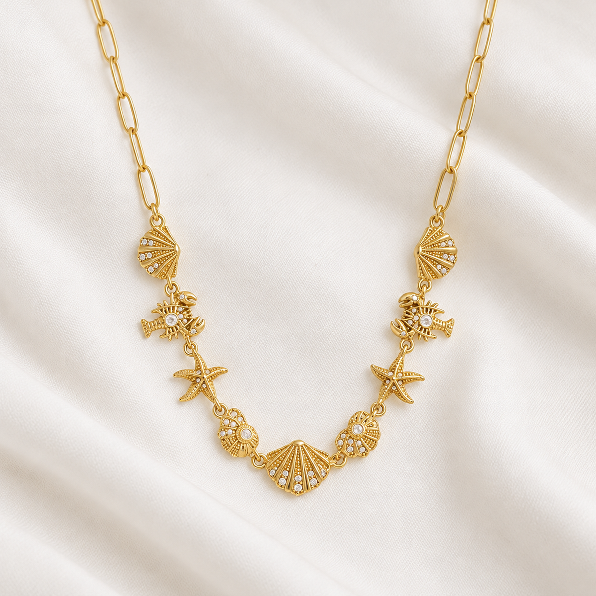 A gold-toned paperclip chain necklace featuring shell, starfish, and sea urchin charms embellished with small clear rhinestones, displayed on a draped cream satin fabric.