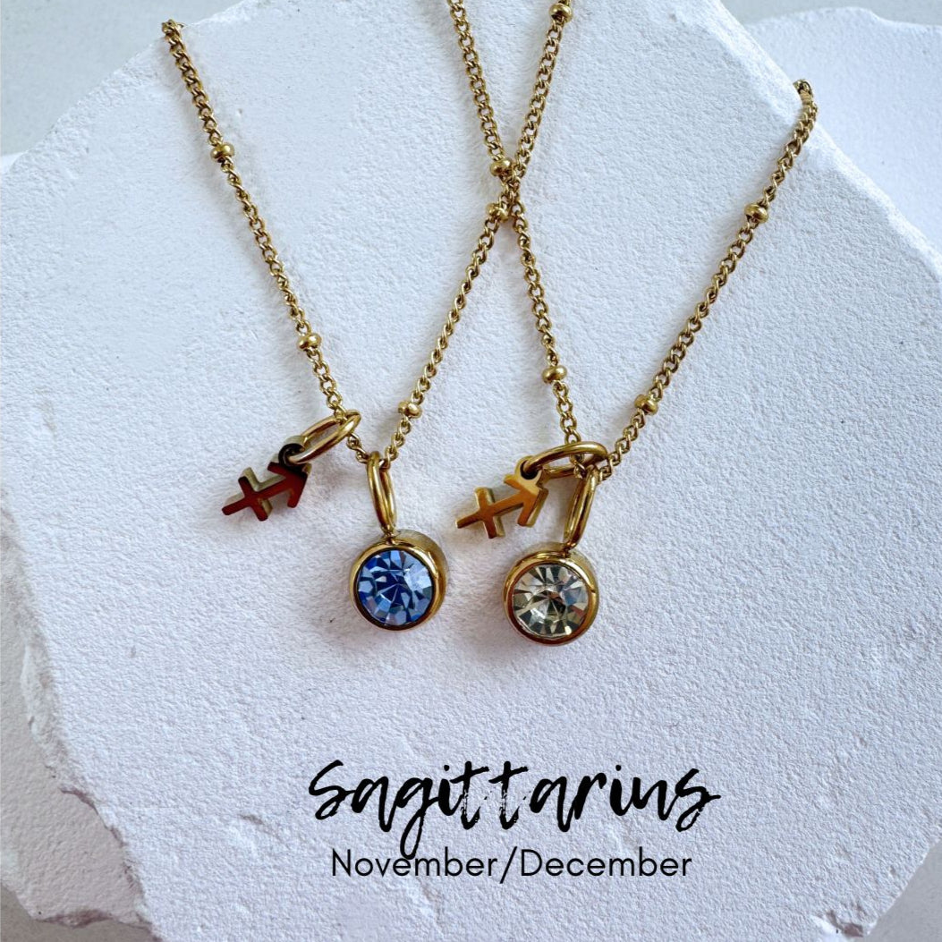 Two gold necklaces with zodiac charm pendants on a textured white surface.