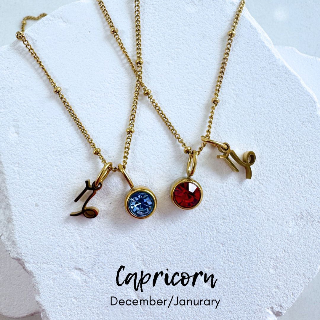 Two gold necklaces with gemstones on a white stone surface, labeled 'Capricorn' and 'December/January'.